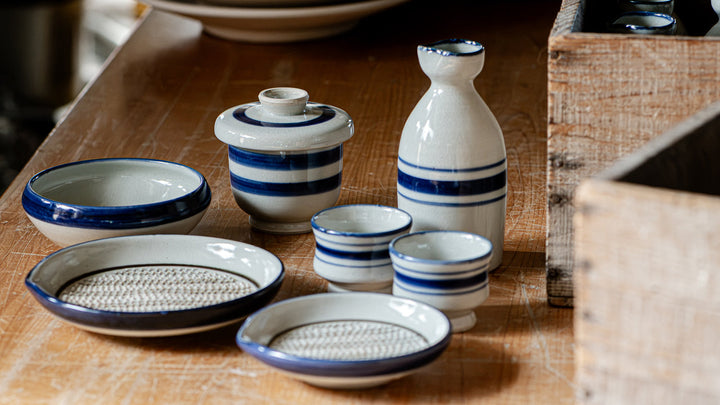 Blue-striped Omi Shimoda ware bowls, a sake bottle, and a grater plate arranged on a wooden table.