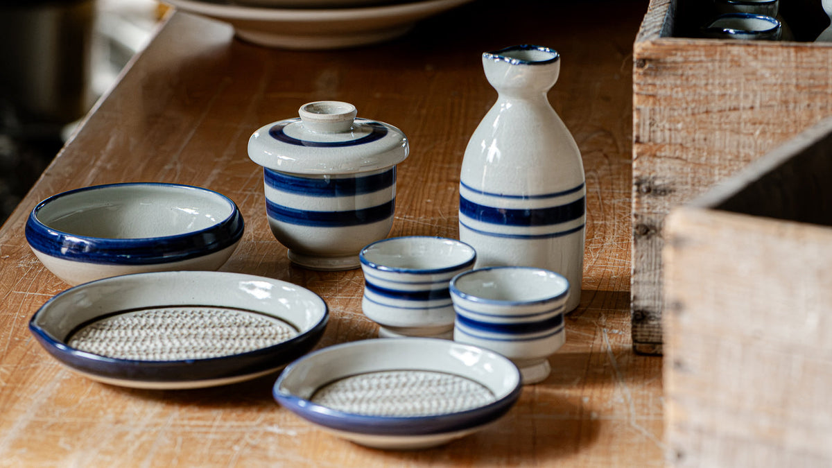 Blue-striped Omi Shimoda ware bowls, a sake bottle, and a grater plate arranged on a wooden table.